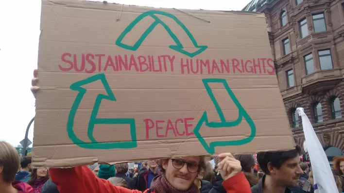 Person mit Schild mit der Aufschrift "Sustainability > Human Rights > Peace" auf einer Demonstration